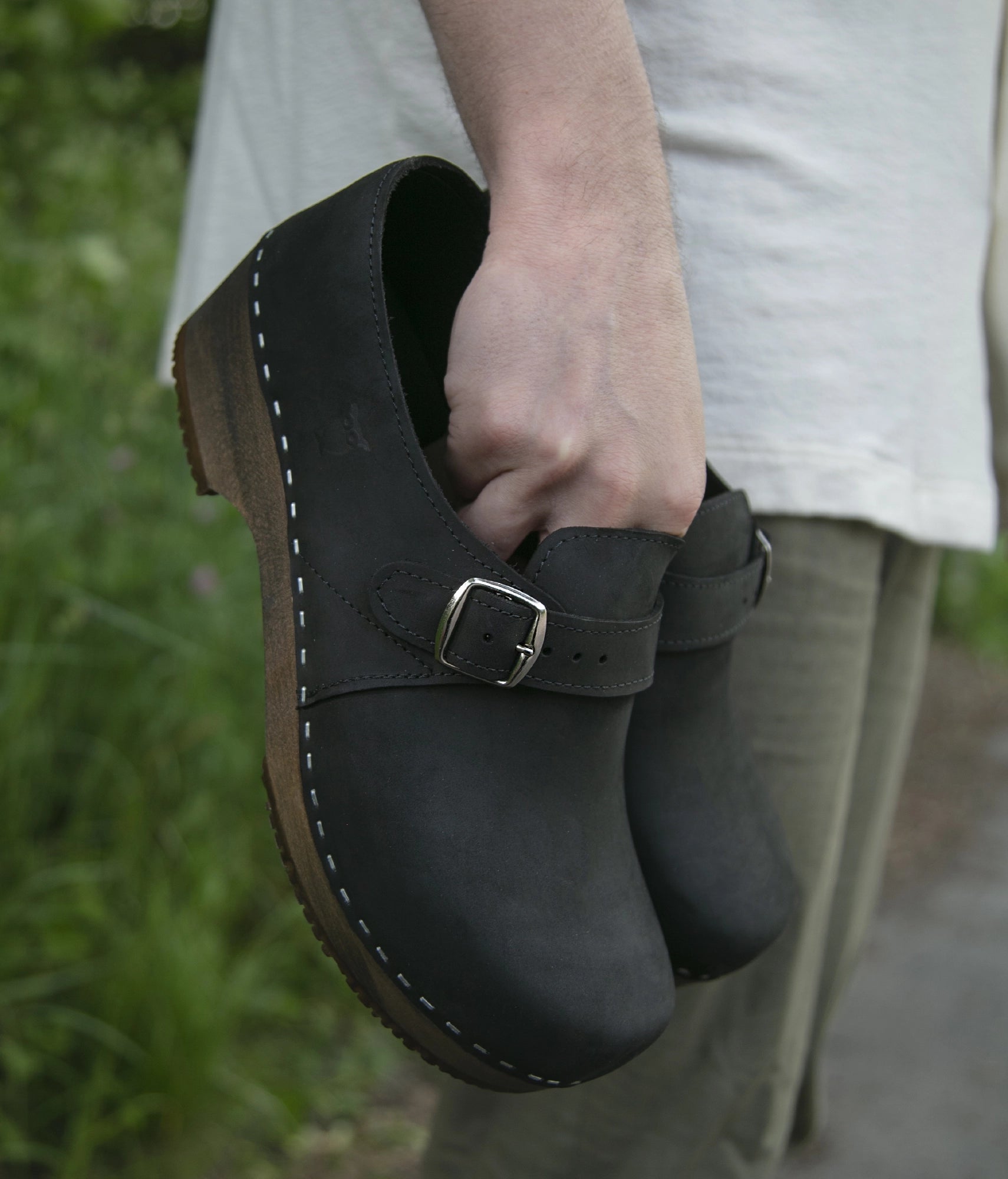 Person holding a black leatherclosed+back clogs with a strap and buckle. men's closed-back clogs in black nubuck leather stapled on a dark wooden base with a silver buckle