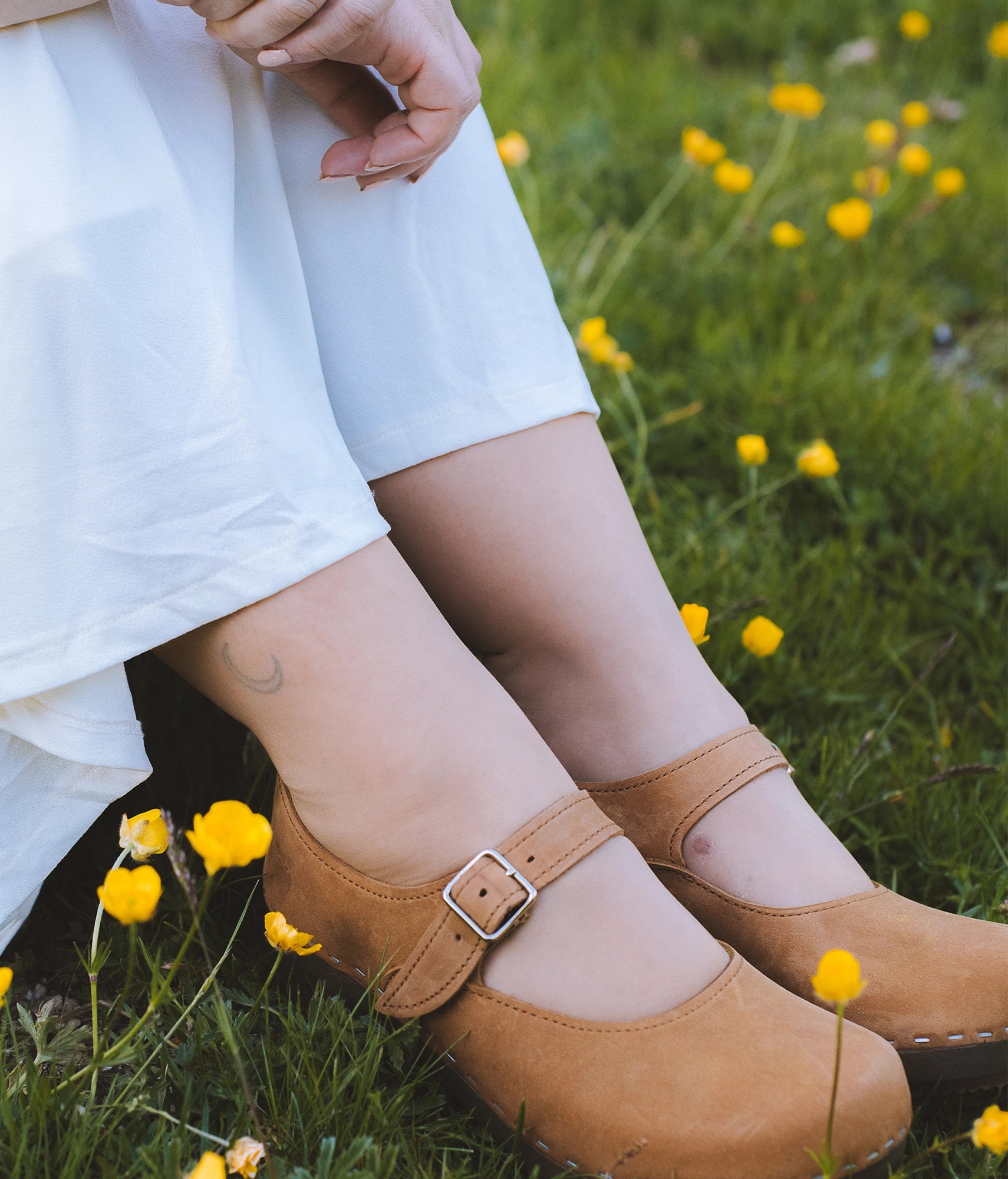 Person wearing Dexter tan closed back clogs with a strap in a grassy field with yellow flowers. Mary Jane wooden clogs in light brown nubuck leather stapled on a dark wooden base