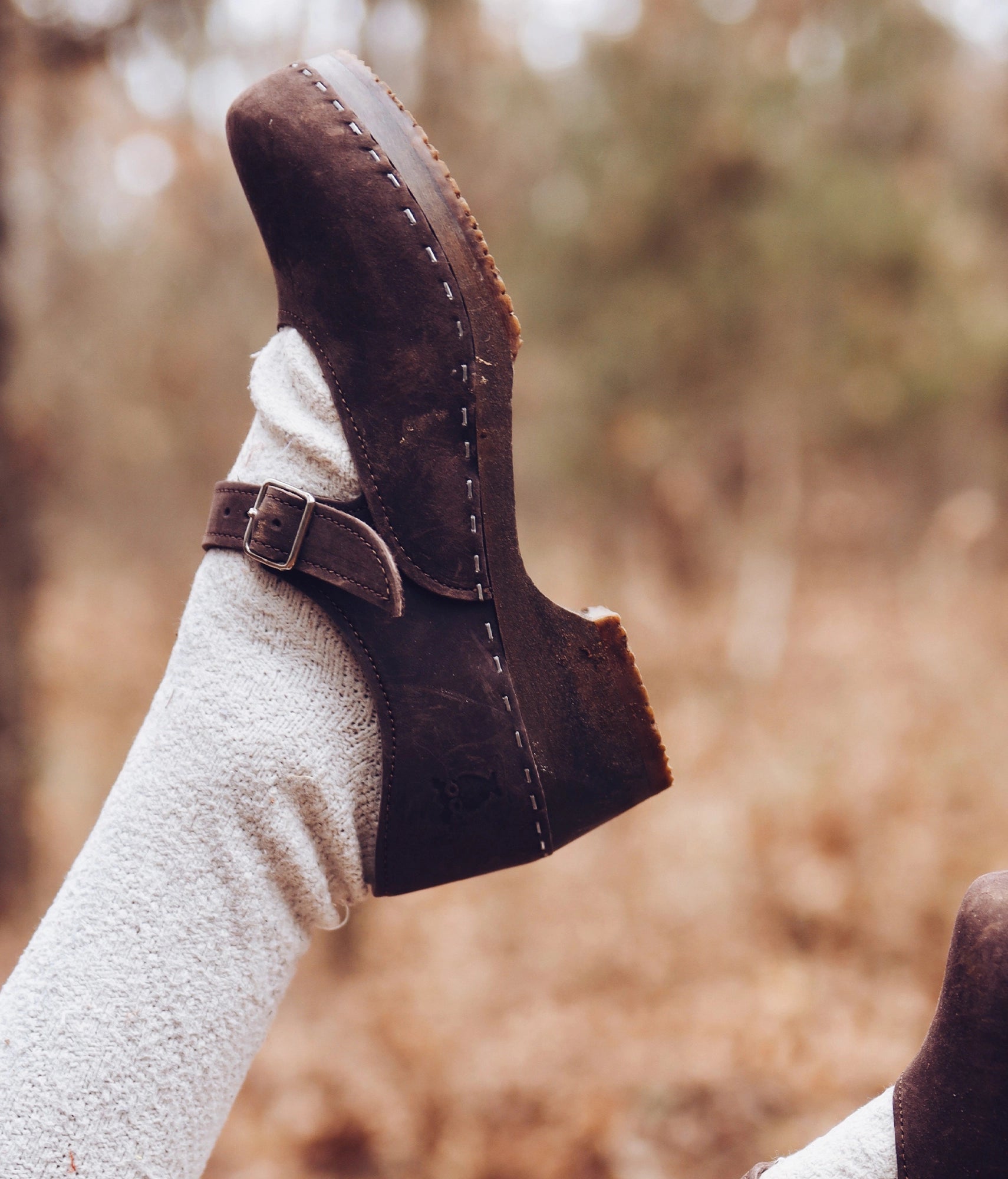 Fudge colored closed-back clogs with a blurred natural background. Mary Jane wooden clogs in dark brown nubuck leather stapled on a dark wooden base