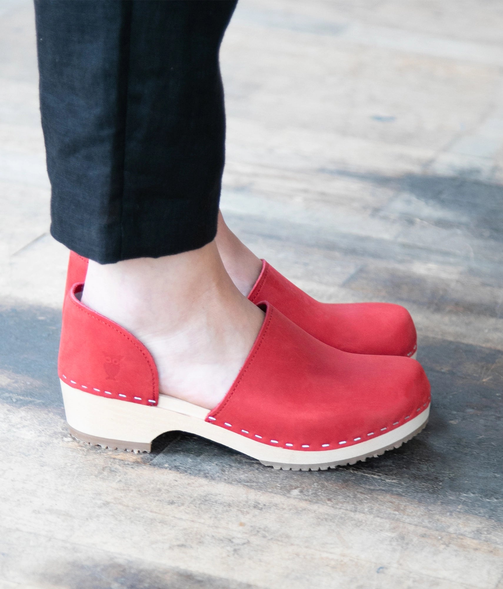 Red closed-back clogs on a light gray background. Low heeled closed-back clogs in red nubuck leather stapled on a light wooden base