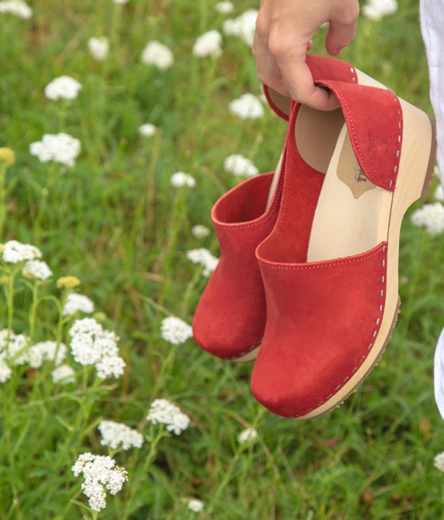Red clogs held above a field of white flowers. Low heeled closed-back clogs in red nubuck leather stapled on a light wooden base