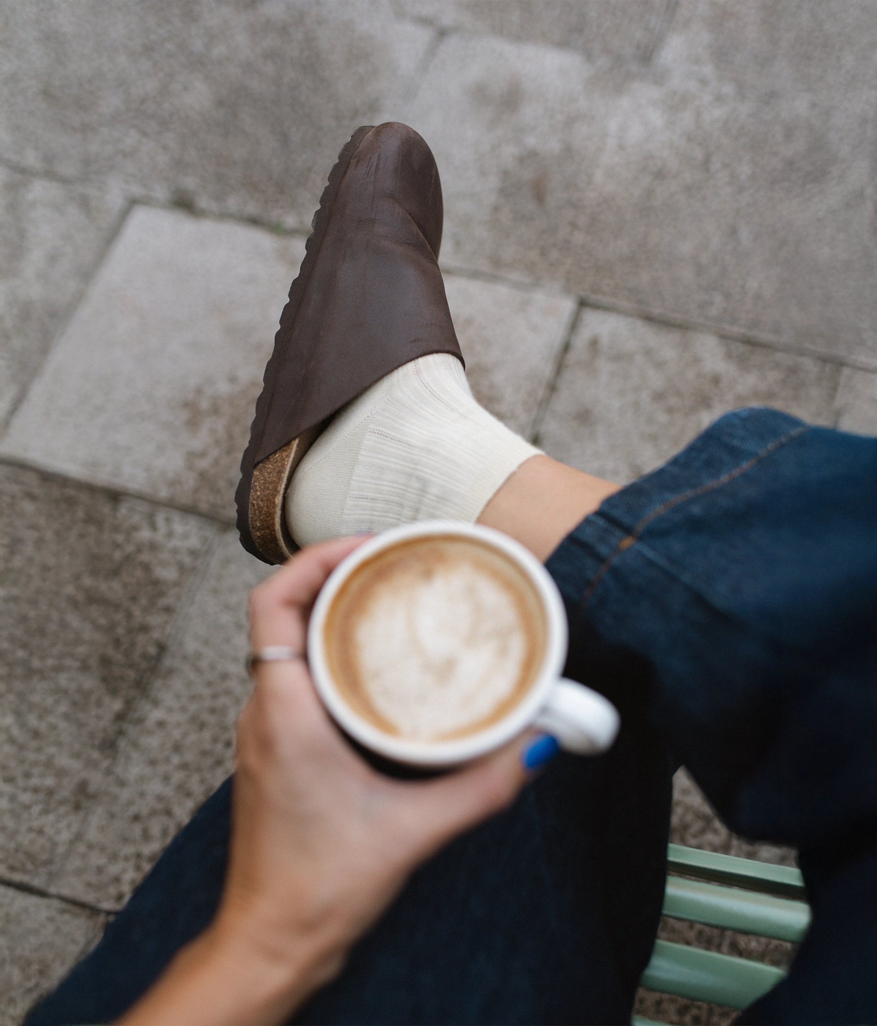 Person holding a cup of coffee with a cork sandals  propped up on a stone pavement
