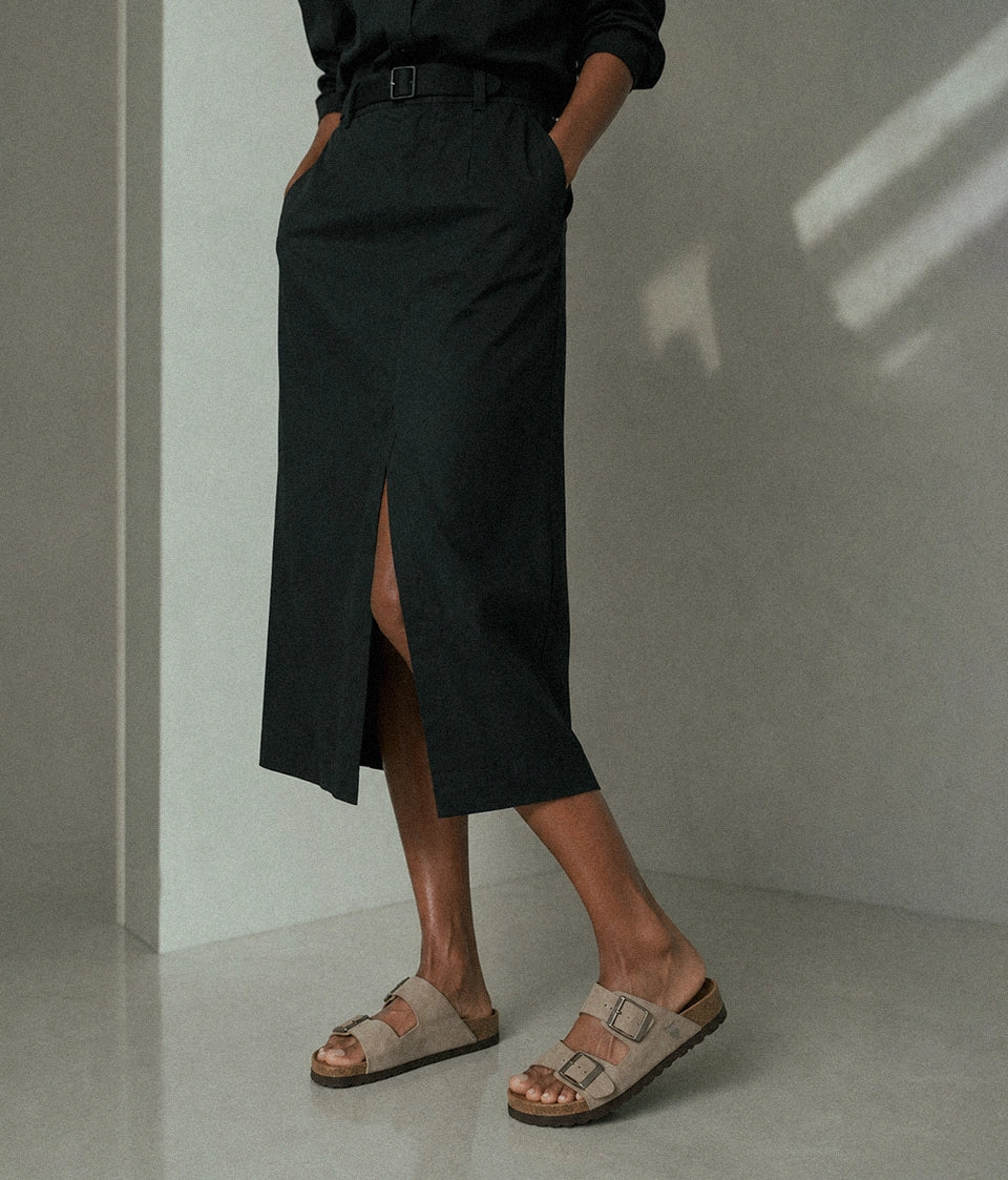 A person wearing a long black midi skirt with a front slit, paired with a black top and beige double-strap slide sandals, standing in a minimalist gray room.
