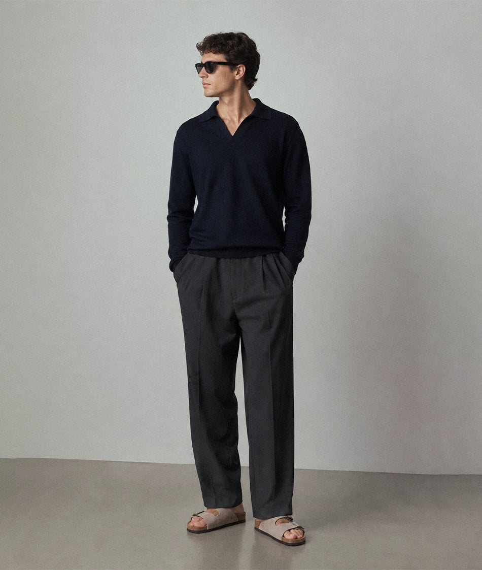 A man wearing a dark navy V-neck knit sweater, dark charcoal gray trousers, and beige cork sandals, standing against a neutral light gray studio background.