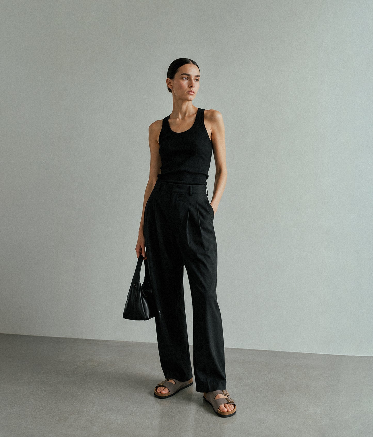 A woman stands in a minimalist studio wearing a black sleeveless top and black wide-leg trousers, holding a black handbag and wearing brown sandals.