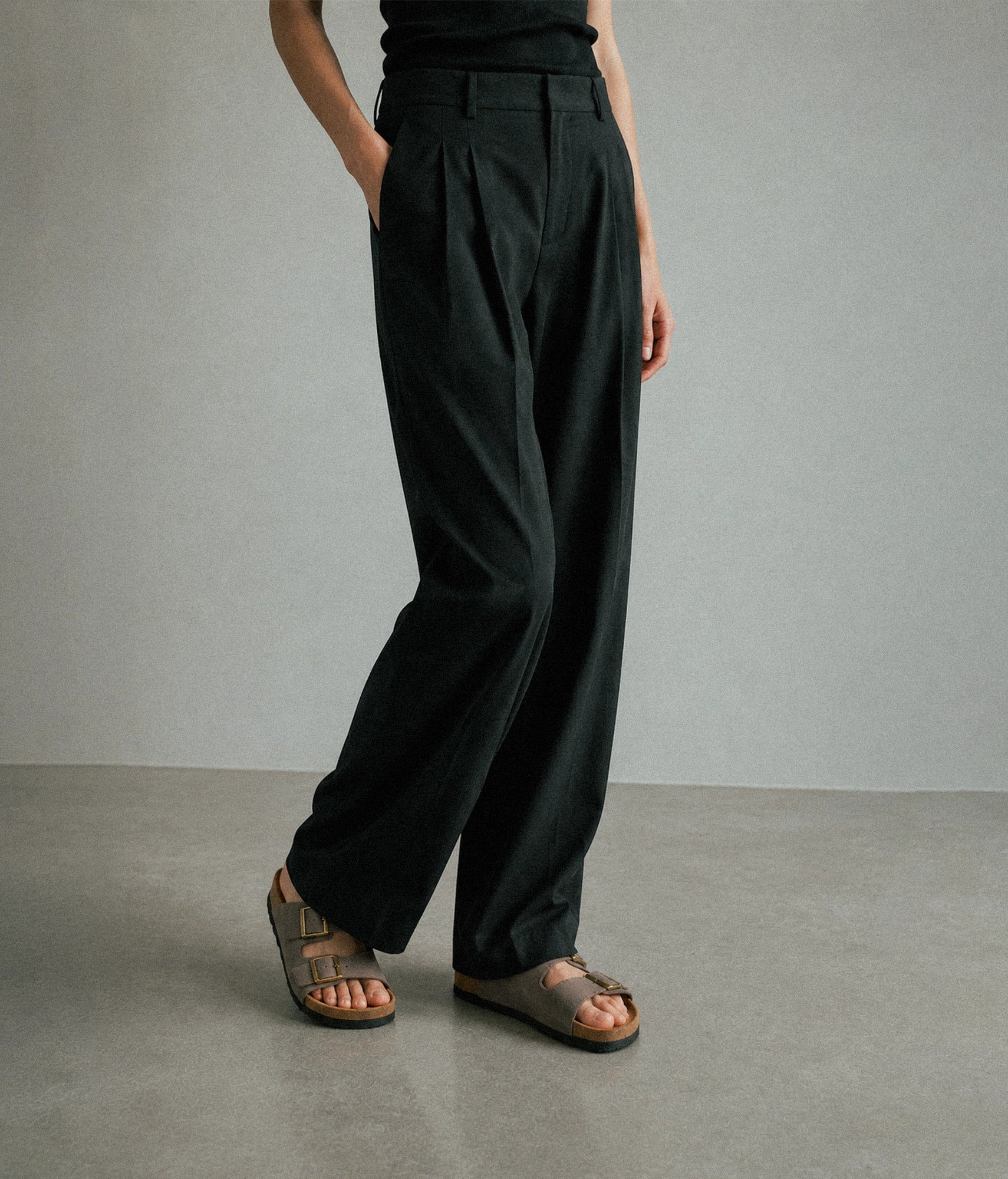 A person wearing black wide-leg pleated trousers with a high waist, styled with brown slide sandals against a neutral gray studio background.
