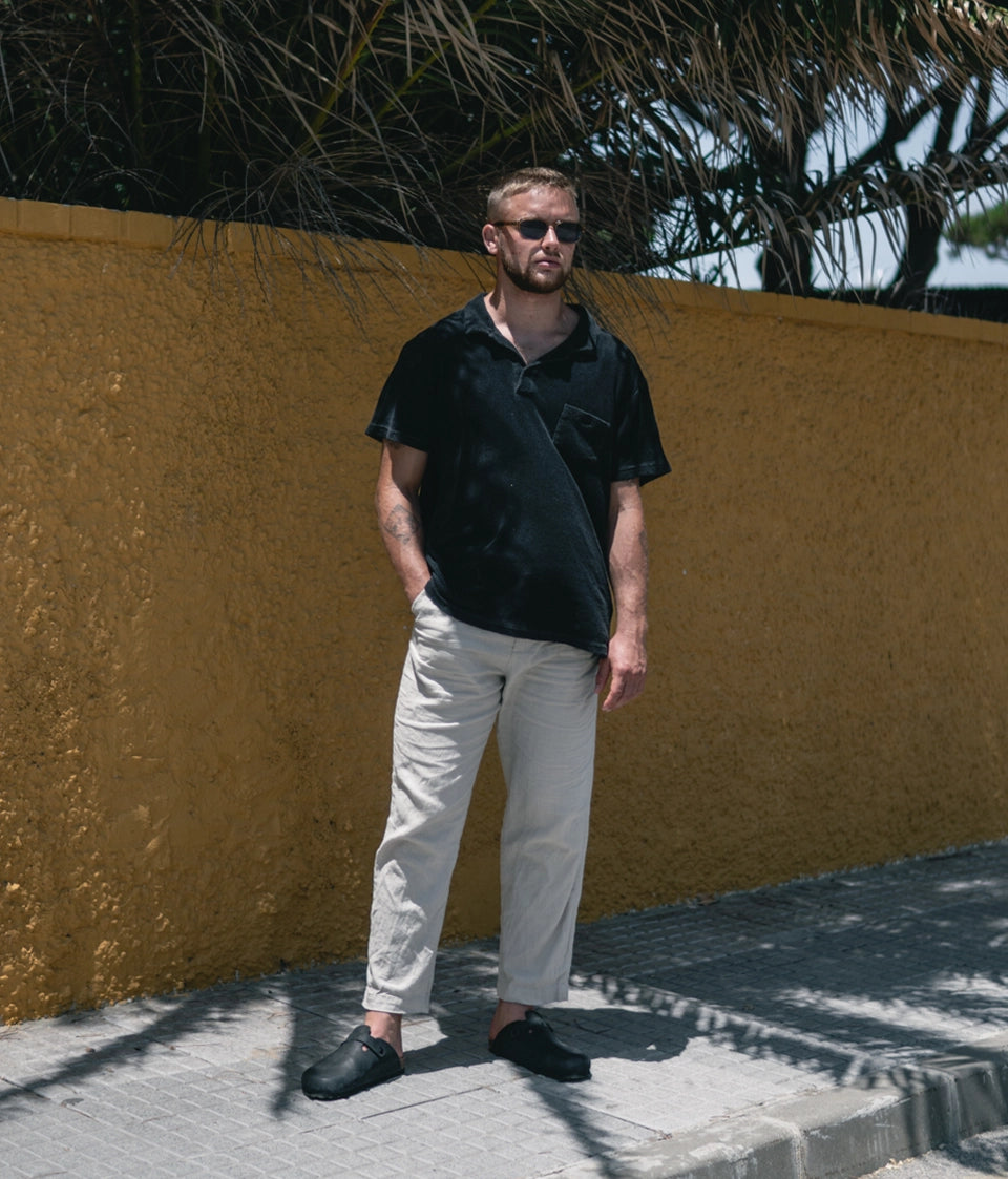 Man wearing sunglasses, a black shirt  and cork mules next to a yellow wall 