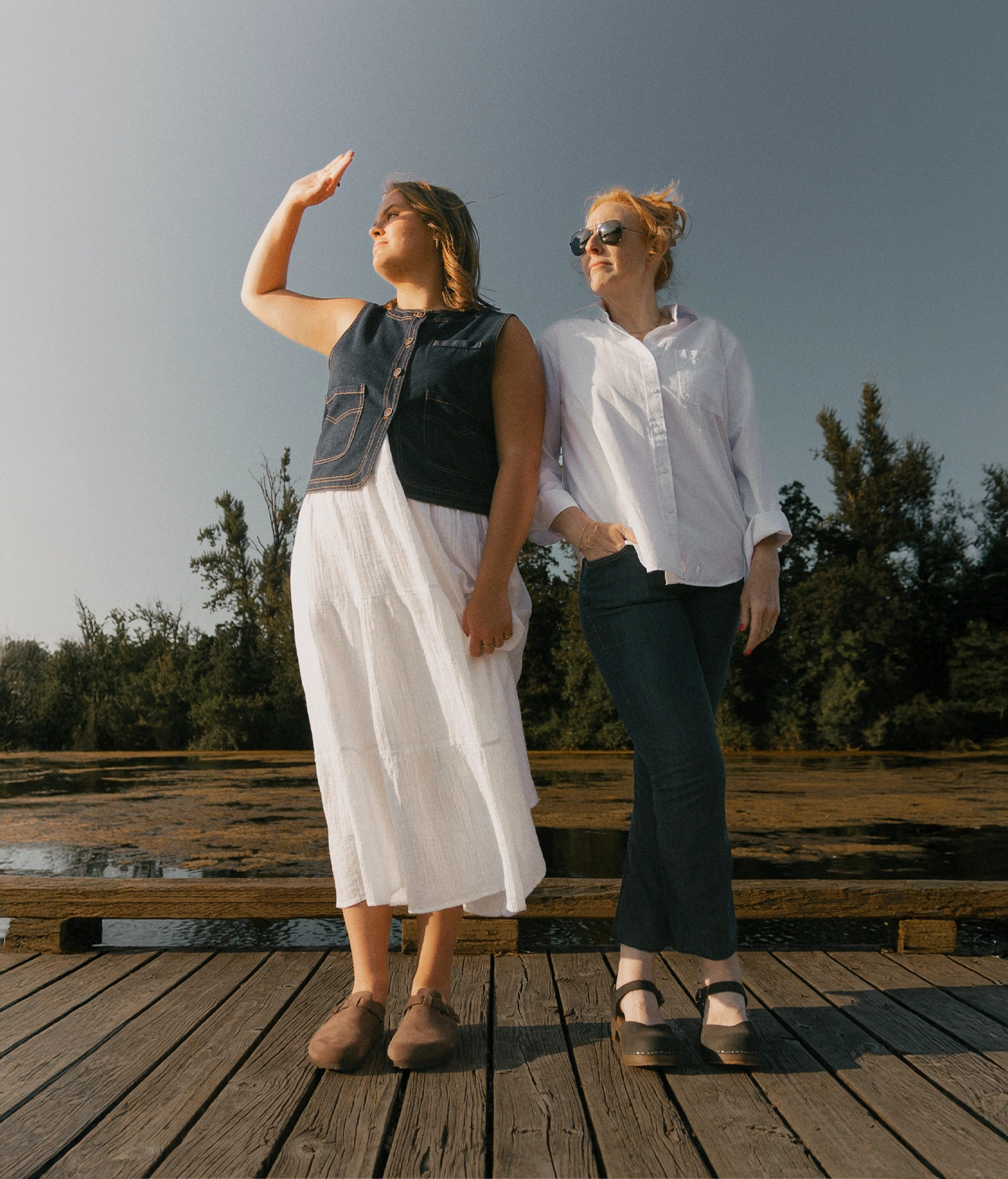 Two women standing on a bridge with Sandgrens footwear on. Classic mules with dark leather. One pair of wooden clogs and another pair of cork mules 